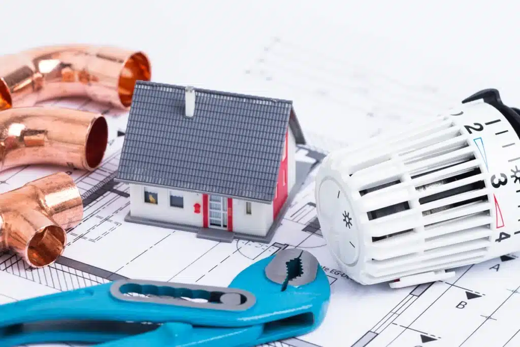 Chauffage et Plomberie : Plan de Rénovation Maison Miniature de maison, raccords cuivre, pince bleue et thermostat sur plans de construction pour projet de chauffage.