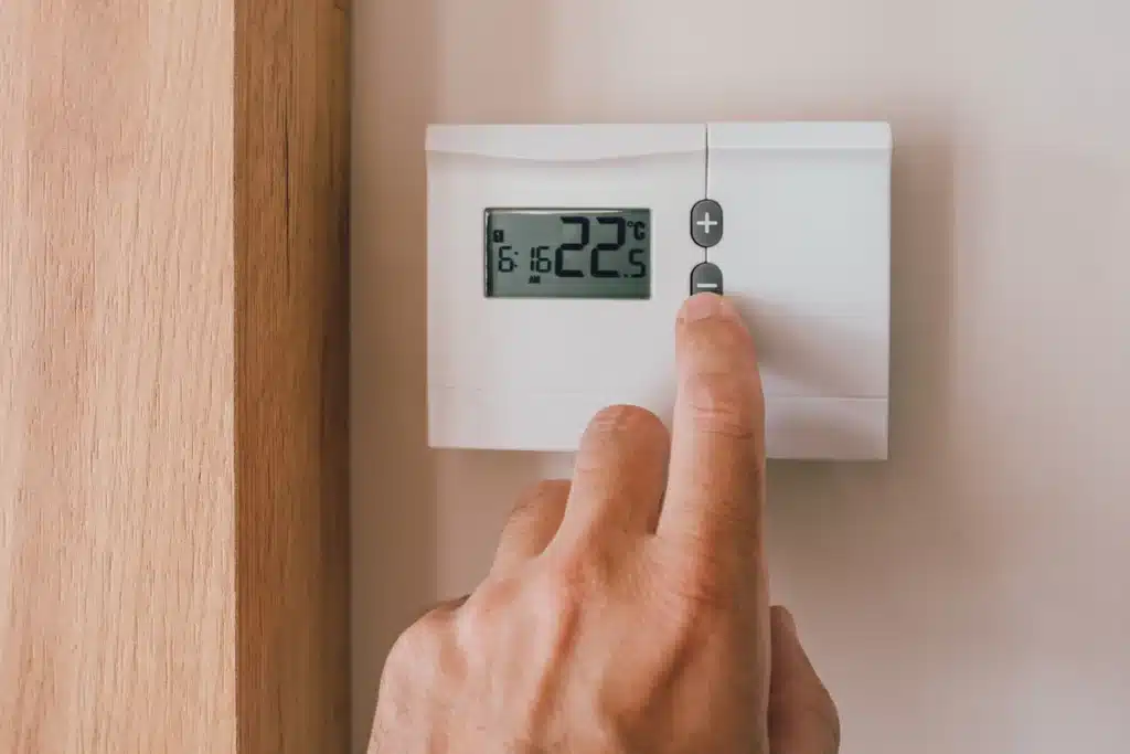Réglage du Thermostat pour Économiser de l'Énergie Main réglant la température à 22.5°C sur un thermostat mural blanc à 6:16 AM pour réduire sa consommation.